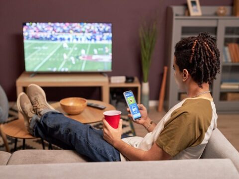 young multicultural male sits at home watching American football on TV while placing a wager through a sports betting app on his smartphone