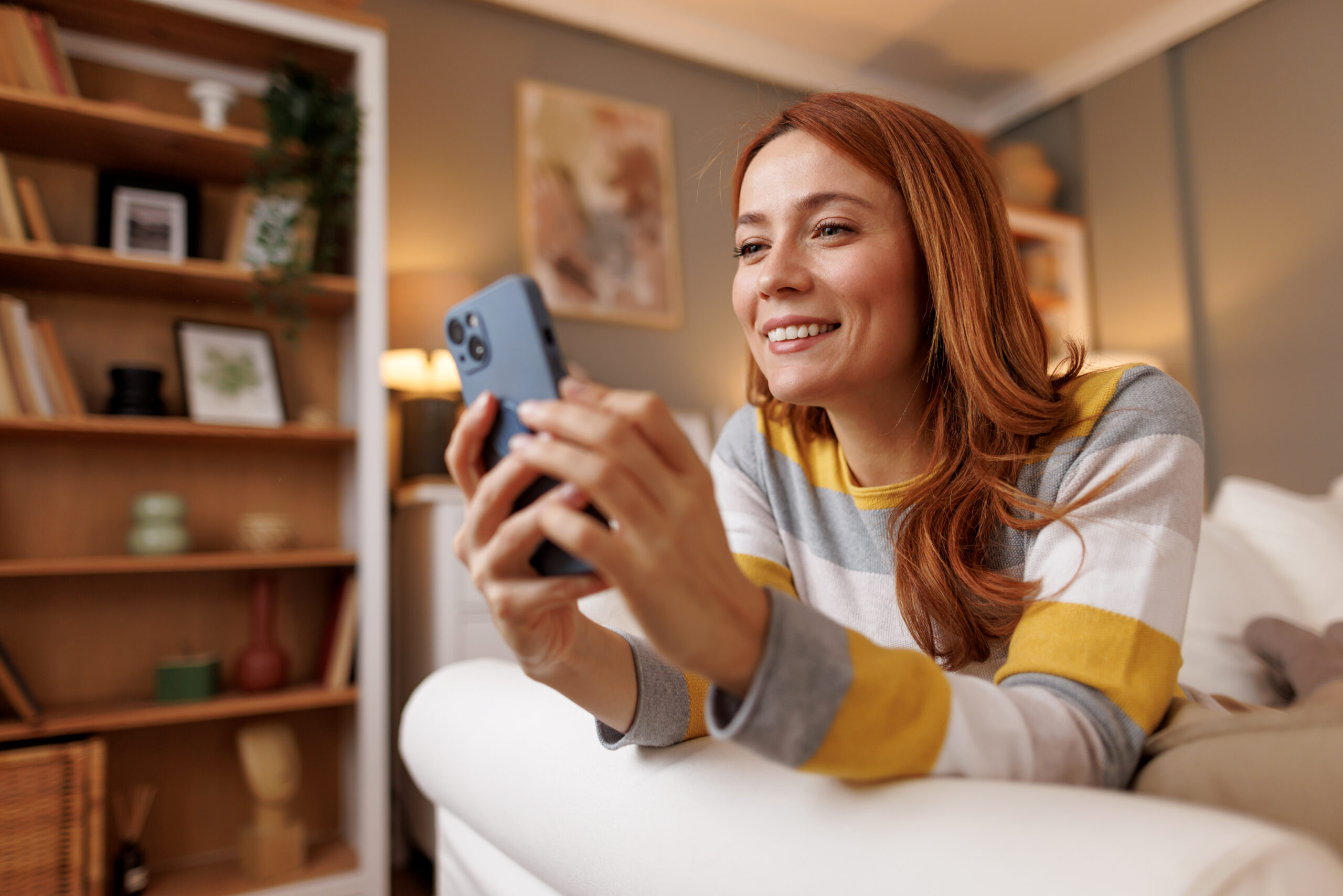 Happy young woman using smartphone while lying on sofa at home