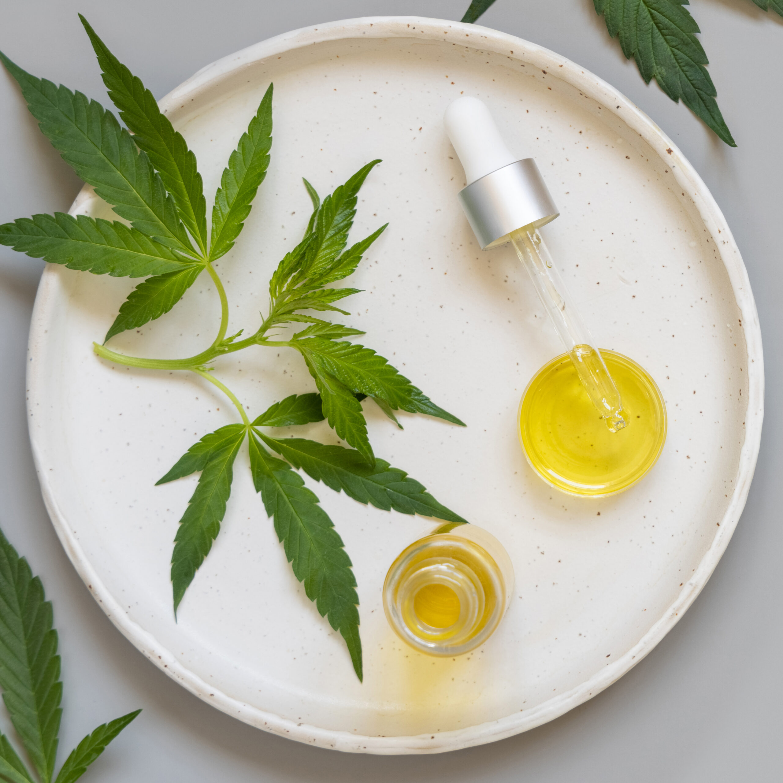 Dropper bottle and pipette near green cannabis leaves on grey ta